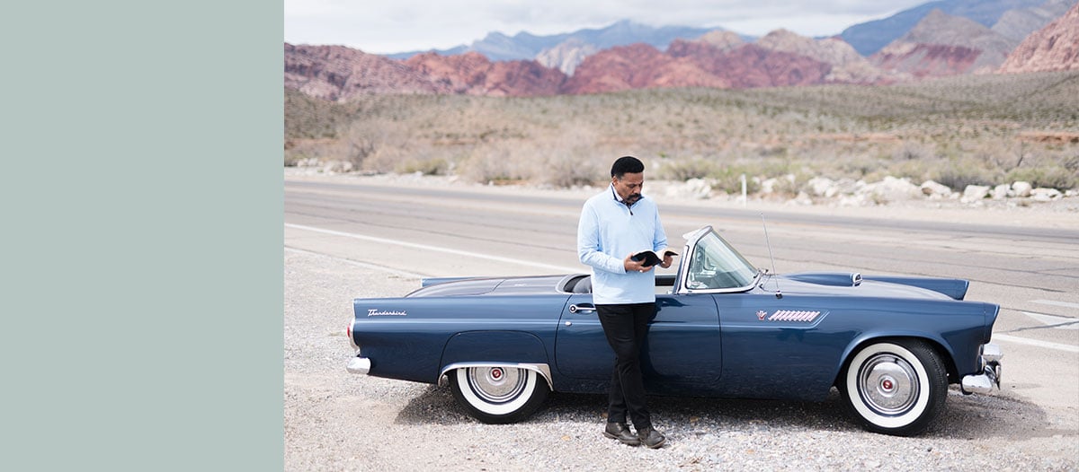Tony Evans standing in front of classic car reading the Bible with desert and mountains in the background.